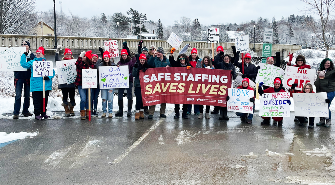 Group photo of RNs and techs by the rally line along the Bad Falls bridge, Machias. Many signs, large banner "Safe staffing saves lives"
