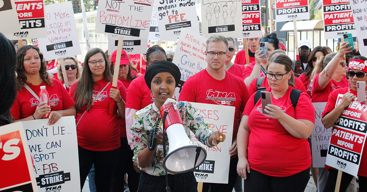 Rep. Ilhan Omar holding megaphone with large group of nurses standing behind her hold strike signs 