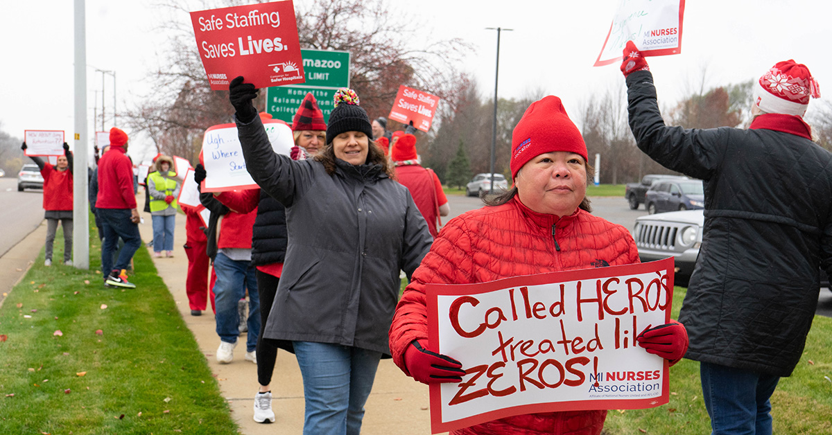 Nurses outside walk line holding signs calling for safe staffing and other demands.