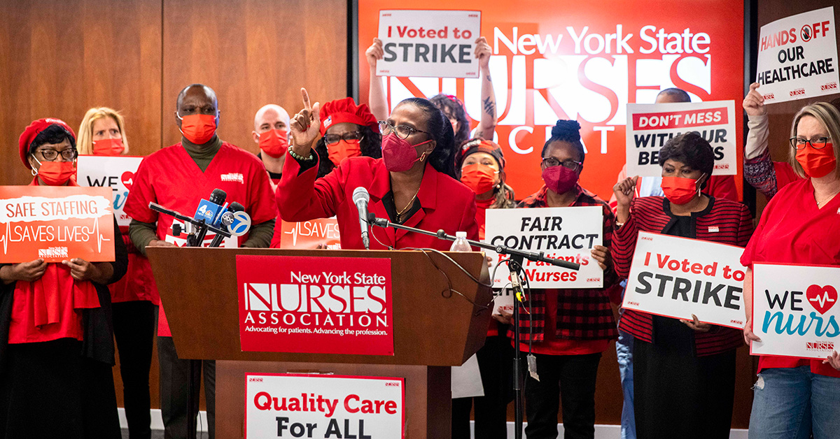Nancy Hagain stands at podium in fron tof large group of NYSNA nurses holding signs "I voted to strike"