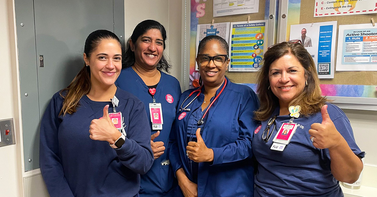 Four nurses inside hospital in blue scrubs giving thumbs up.