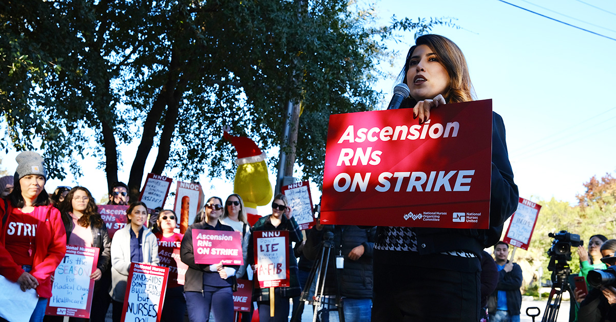 Nurse speaking into microphone holding sign "Ascension RNs On Strike". Other nurses with signs behind.