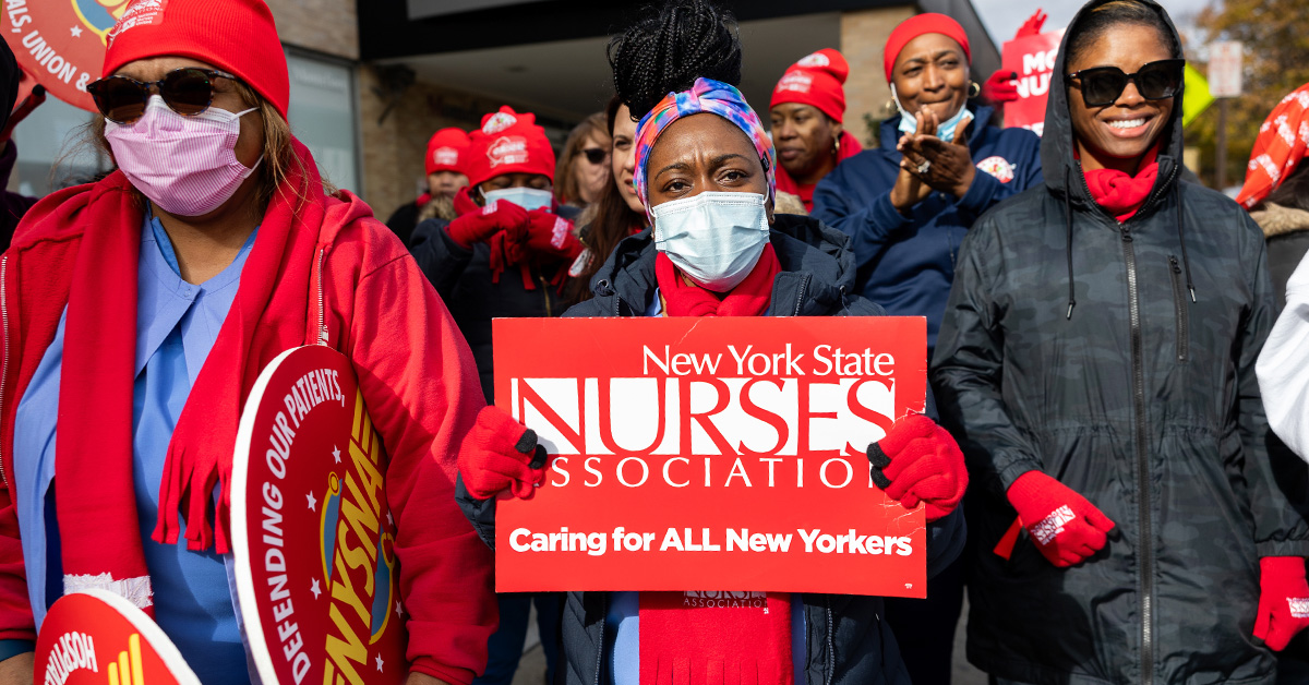Group of nurses walking out of hospital together, one holds sign "New York State Nurses Association: Caring For All New Yorkers"