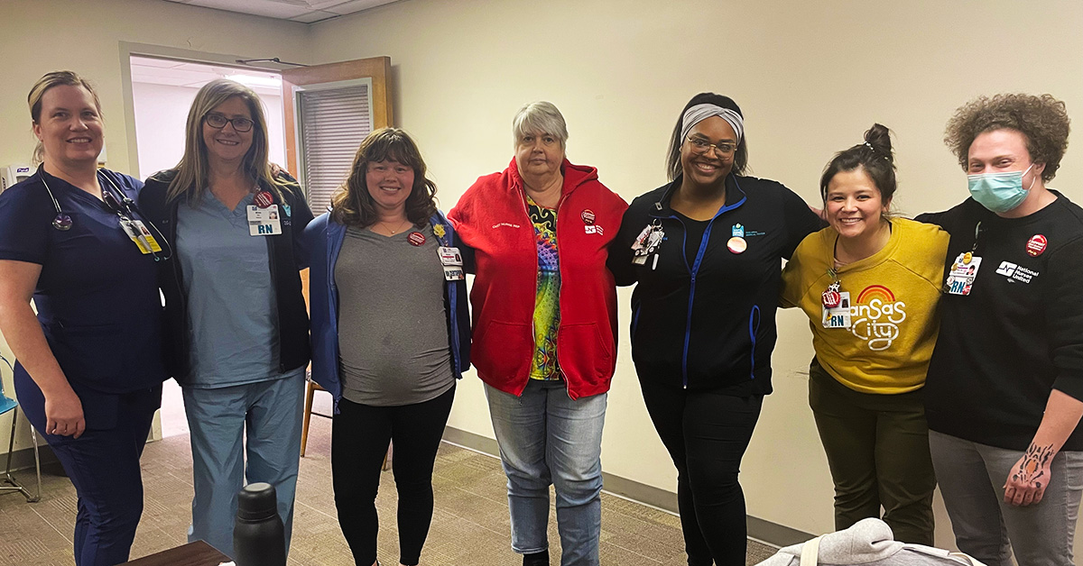 Group of seven nurses inside hospital smiling with arms around each other