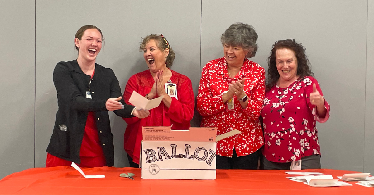 Four nurses standing at table with ballot box, smiling and laughing