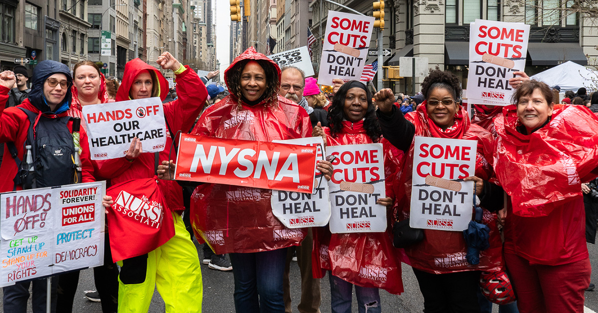 Group of nurses outside in rain clothes holding signs "Some Cuts Don't Heal"