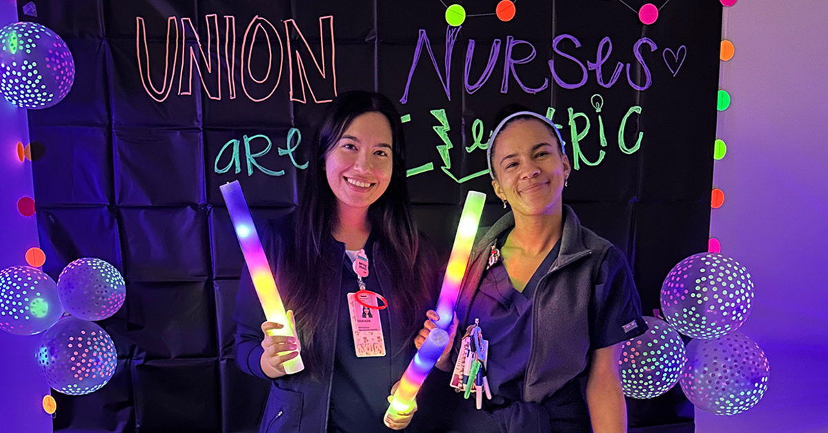 Two nurses smiling and holding glod sticks in front of banner "Union Nurses are Electric"