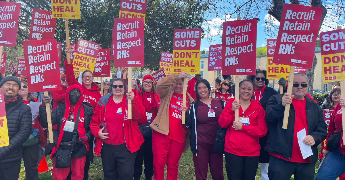 Large group of nurses on picket line, holding signs "Some Cuts Don't Heal" and "Recruit, Respect, Retain RNs"
