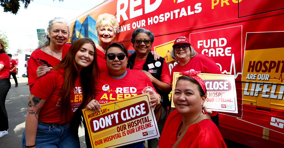 Group of nurses in front of Red Alert tour bus, smiling, holding sign "Don't close our hospital"