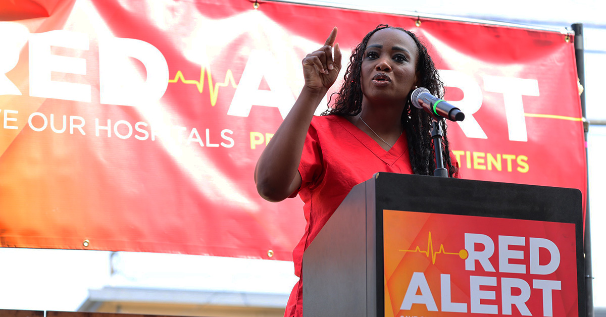 Nurse speaking at podium with banner behind "Red Alert: Save Our Hospitals Tour"