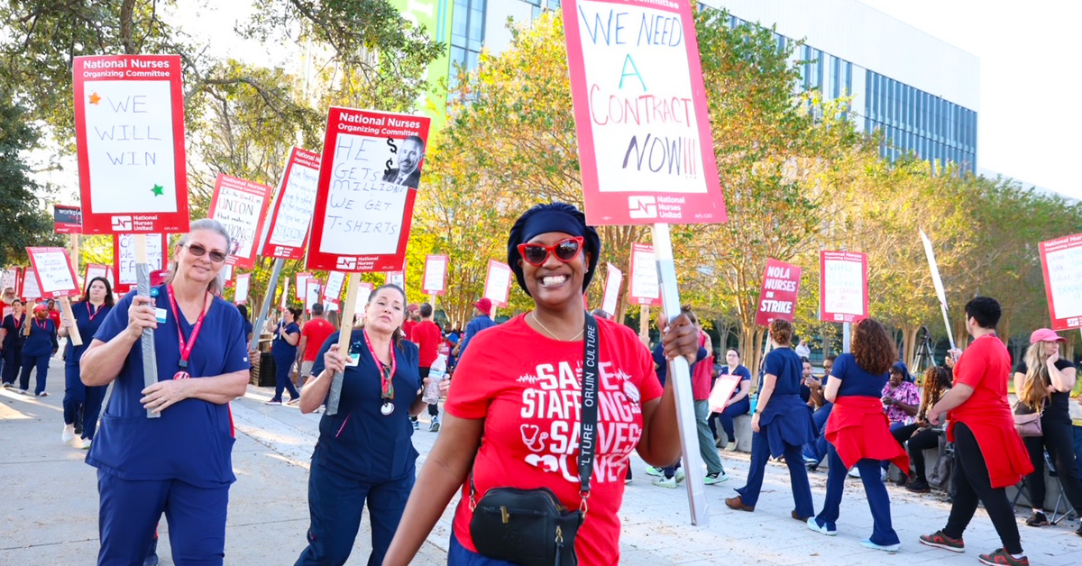 Nurses on picket line, smiling, one holds sign "We Need a Contract NOW!"