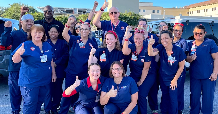 Large group of nurses outside hospital, wearing blud scrubs, smiling, and giving thumbs up