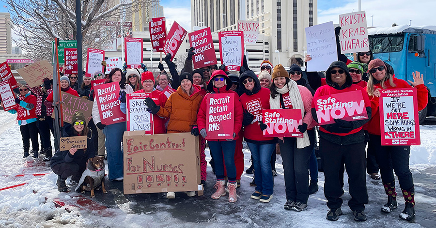 Large group of nurses outside hospital, in cold weather clothes, holding signs including "Safe Staffing NOW" and "Recruit, Retain, Respect RNs"