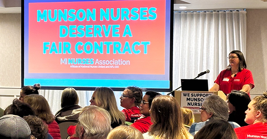 Nurse at podium in town hall meeting, presentation on screen titled "Munson Nurses Deserve a Fair Contract"