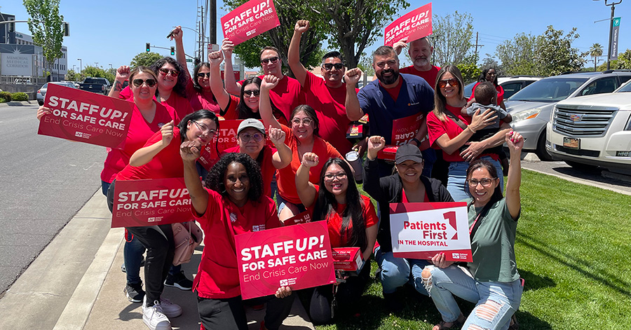 Group of nurses outside, wearing red, smiling, raised fists, holding signs "Staff Up For Safe Patient Care"