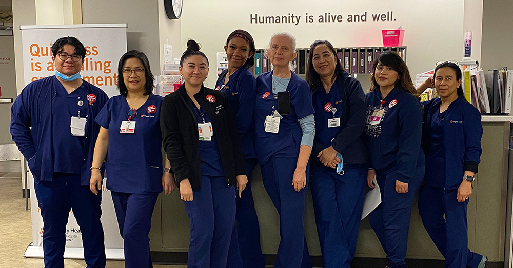 Group of eight nurses inside hospital, side-by-side, in blue scrubs