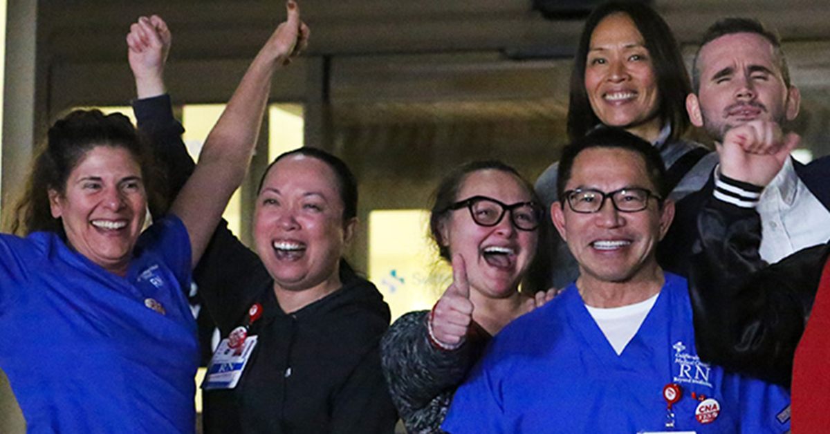 Large group of nurses outside hospital, smiling, with raised fists