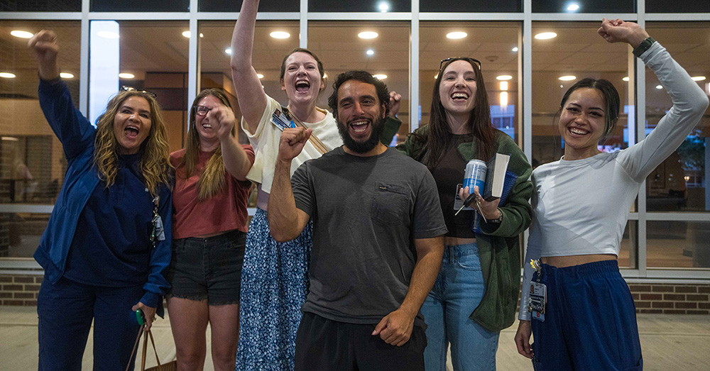 Group of six nurses outside hospital, smiling, with raised fists