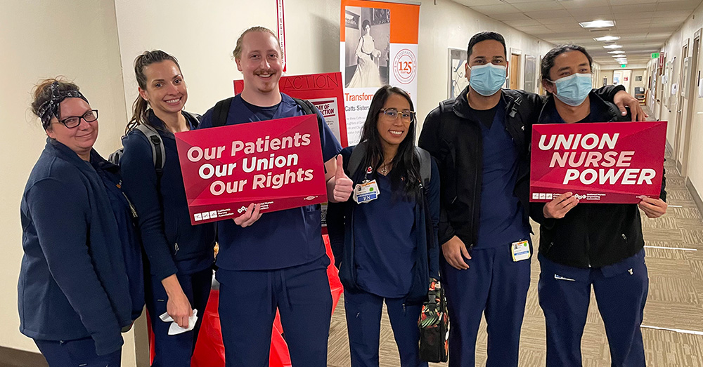 Six nurses inside hospital, side-by-side, smiling, two holding signs "Union Nurse Power"