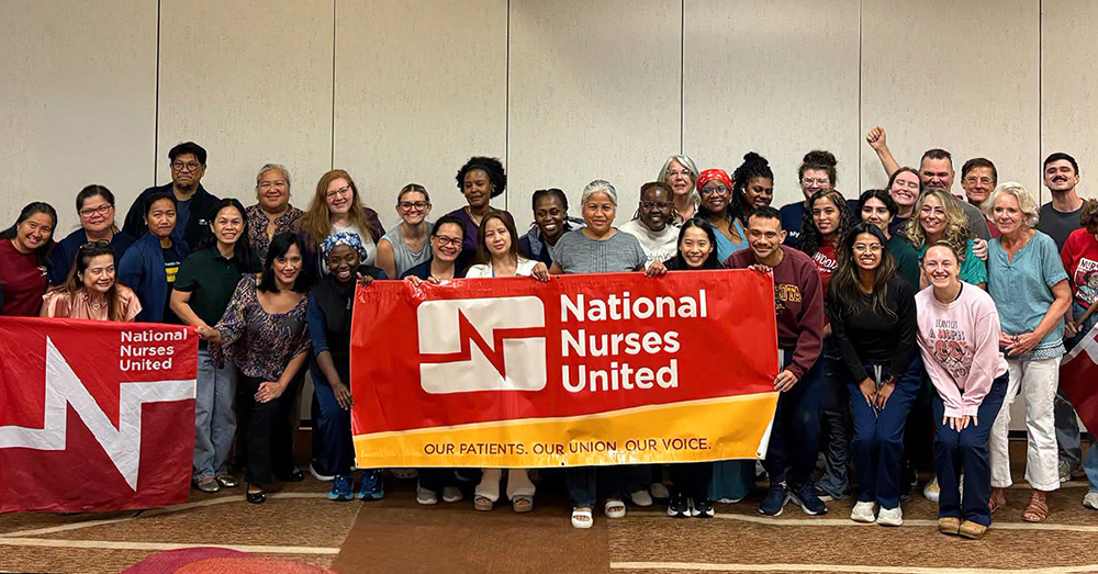 Large group of nurses inside conference room holding banner "National Nurses United"
