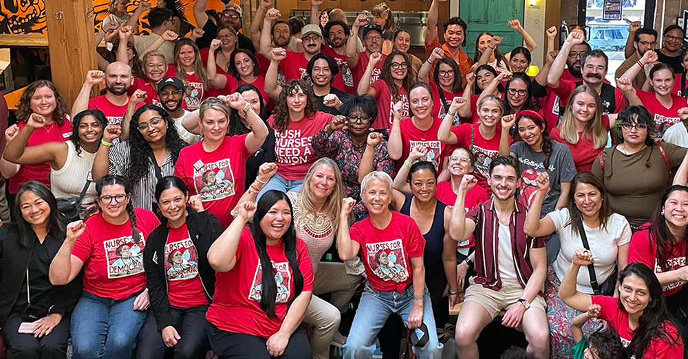 Large group of nurses inside, mostly wearing red, smiling, with raised fists