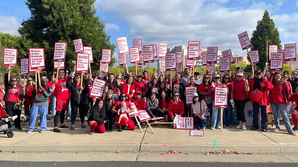 Large group of nurses outside holding signs "Kaiser: Put Patients Over Profit" and "Nurses at the Bedside, Kaiser at the Bank"