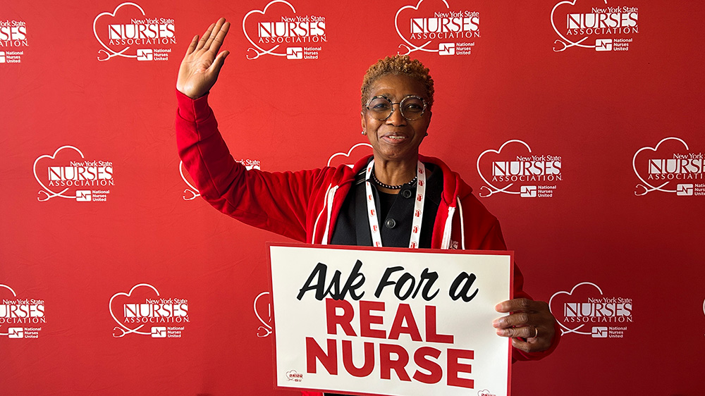 Nurse smiling, with one hand in air, holding sign "Ask for a Real Nurse"