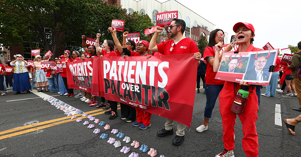 Nurse standing in street holding banner "Patients Over Palantir"