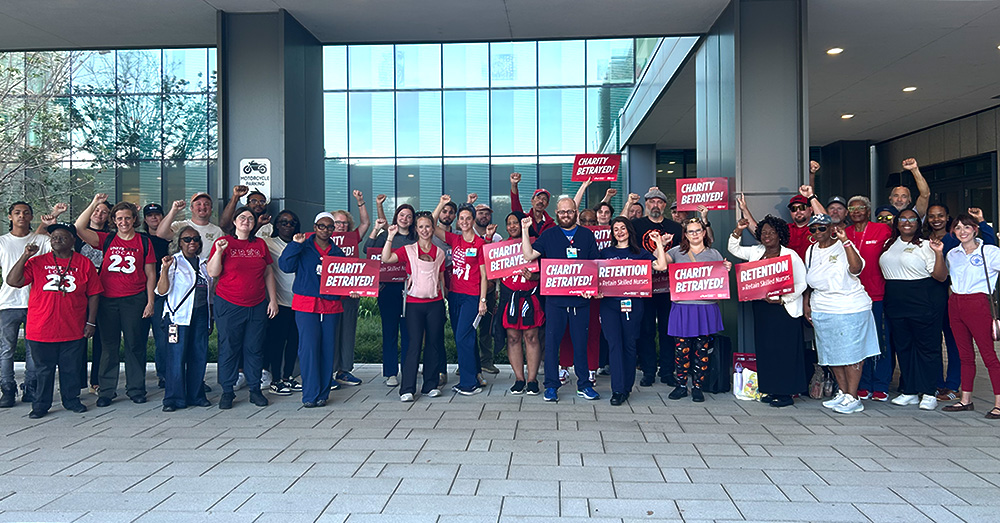 Large group of nurses outside hospital with raised fists, some holding signs "Charity Betrayed"