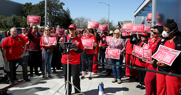 Nurse speaking at podium surrounded by other nurses holding signs "Protect Nurses, Patients, Public Health"