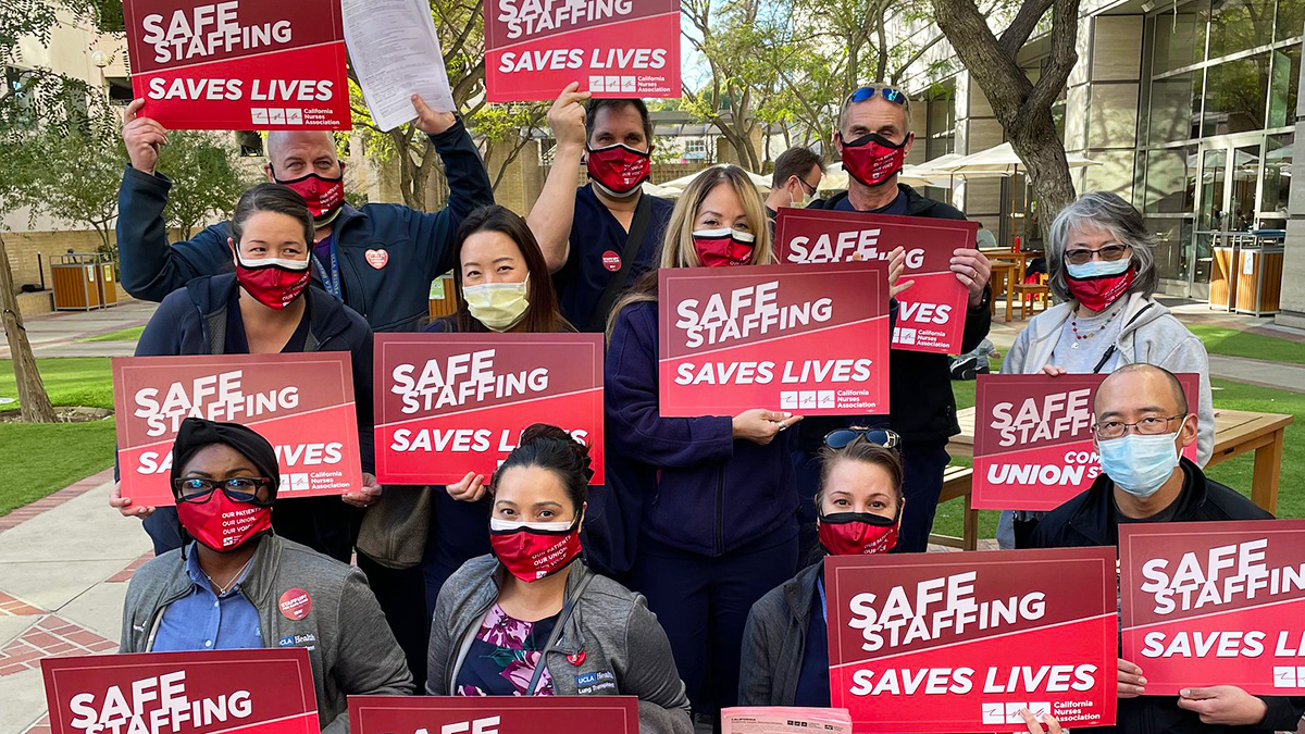 Large group of nurses outside hospital hold signs "Staff Up for Safe Care"