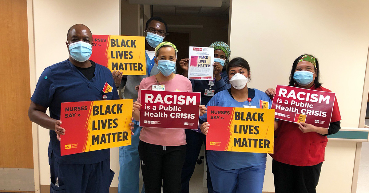 Group of nurses inside hospital hold signs "Nurses Say: Black Lives Matter" and "Racism is a Public Health Crisis"