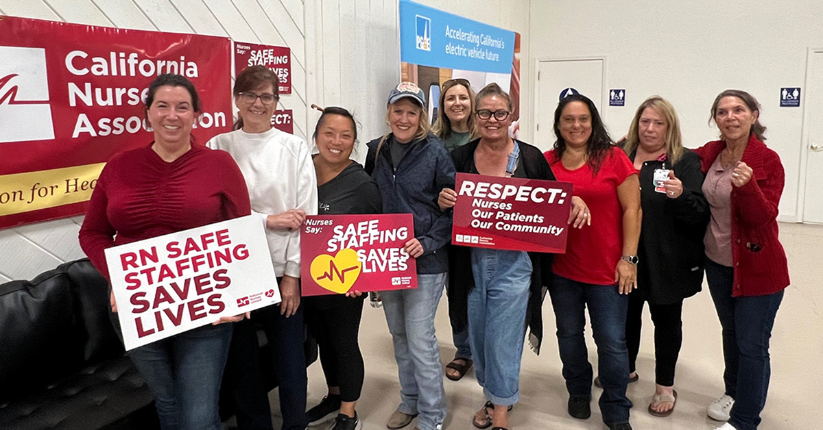 Group of 9 nurses smiling, one holds signs "Respect Nurses, Our Patients, Our Communitites