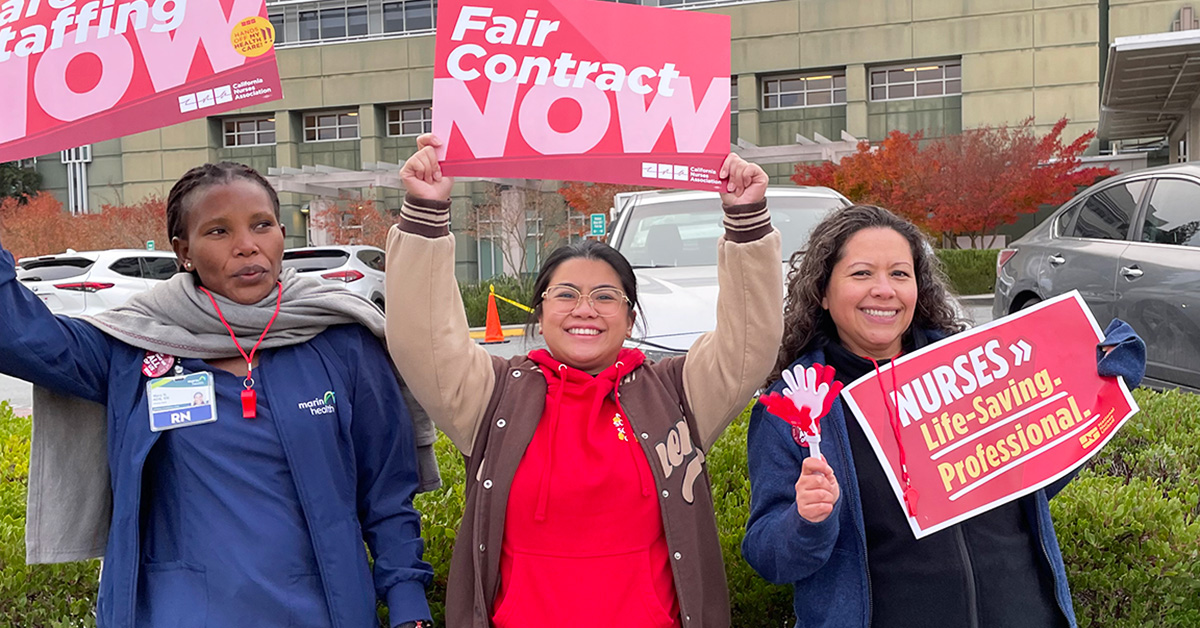 Three nurses outside hospital holding signs "Fair Conract Now" and "Safe Staffing Now"
