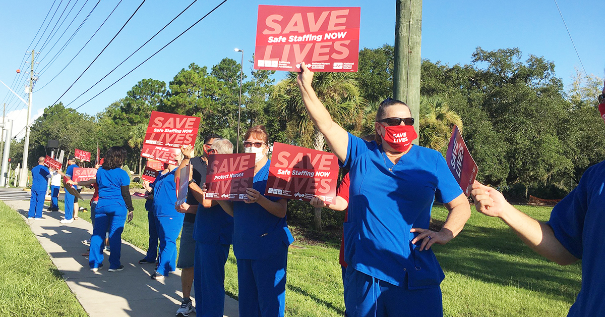 Many nurses holding signs on sidewalk: "Save Lives, Safe Staffing NOW"