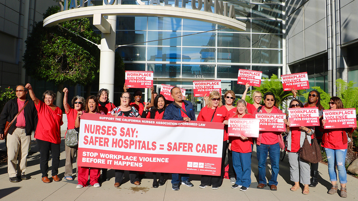 Large groupe of nurses outside hospital hold signs "Safer Hospitals = Safer Care"