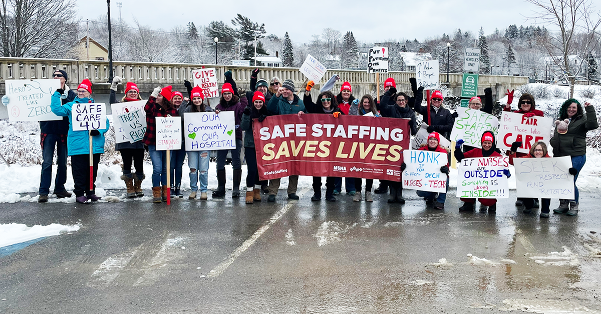 Group photo of RNs and techs by the rally line along the Bad Falls bridge, Machias. Many signs, large banner "Safe staffing saves lives"