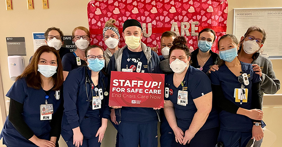 Large group of nurses inside hospital, one holds sign "Staff Up for Safe Care"