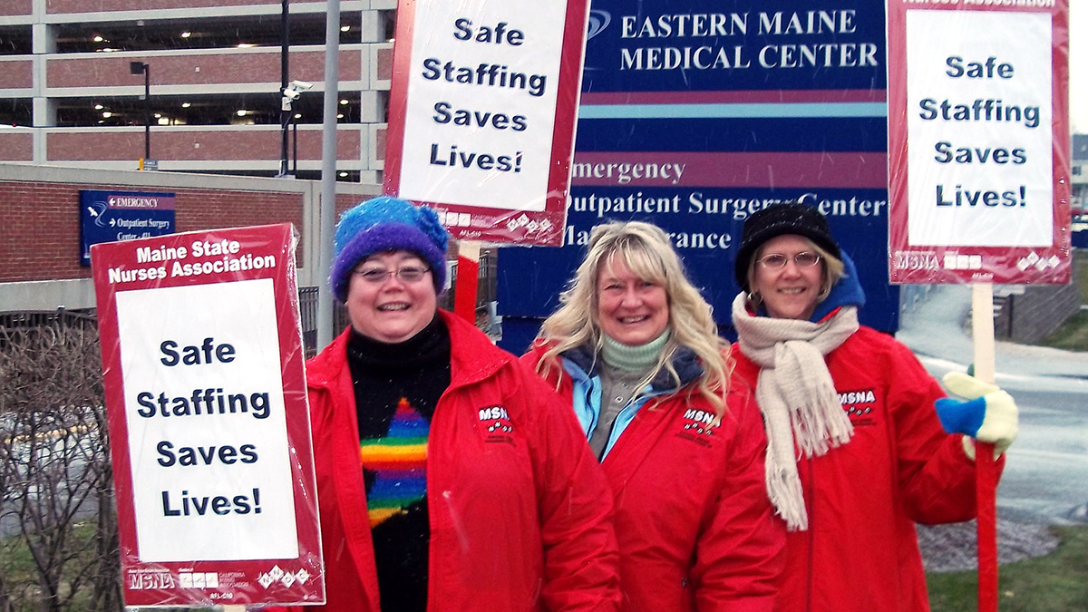 Three nurses outside Eastern Maine Medical Center hold signs "Safe Staffing Saves Lives"