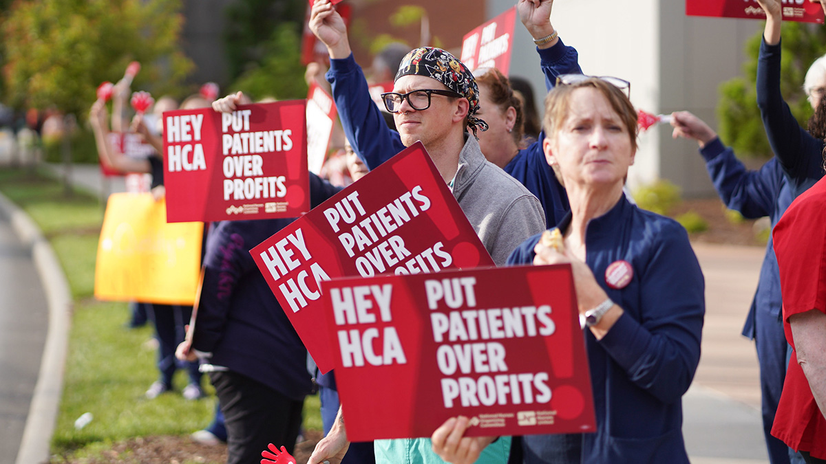 Nurses on picket line outside hospital holding signs "Hey HCA Put Patients Over Profit"