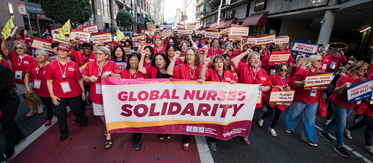 Large group of nurses marching through streets holding banner "Globarl Nurses Solidarity"