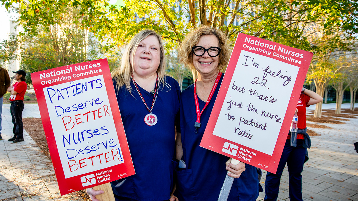 Two nurses smiling and holding signs "Patients Deserve Better, Nurses Deserve Better"