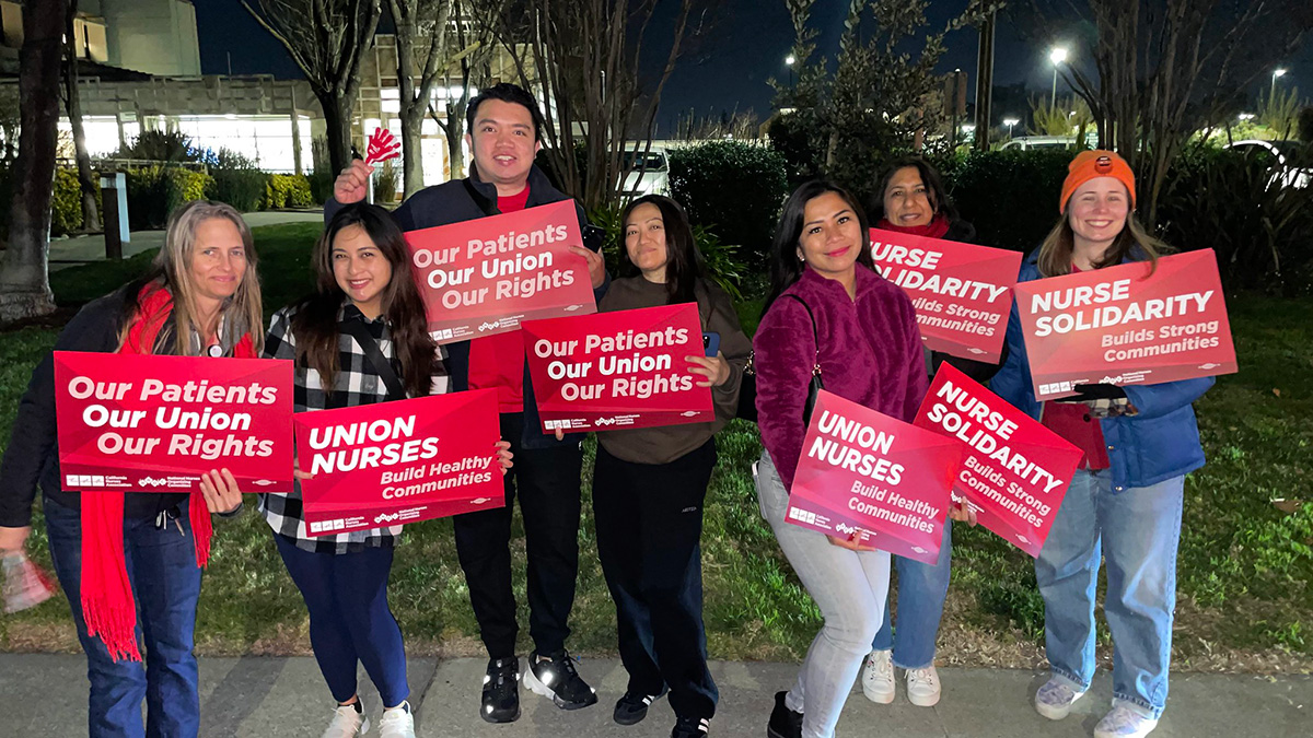 Group of nurses outside hospital at night, holding signs and banner "Our Patients, Our Union, Our Rights"