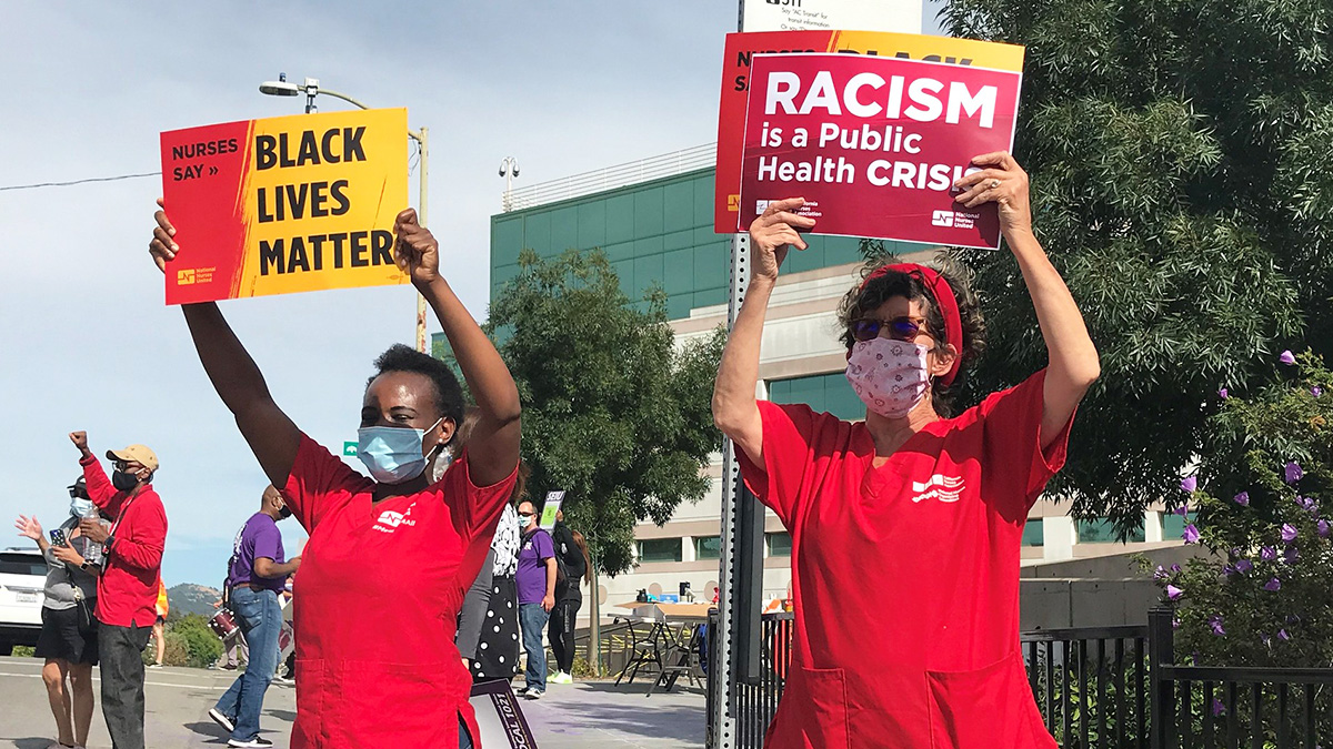 Two nurses outside hold signs "Black Lives Matter" and "Racial Justice is a Public Health Crisis"