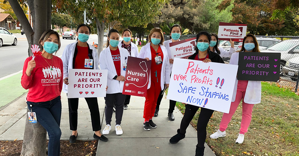 Large group of nurses outside hospital hold signs "Patients First"