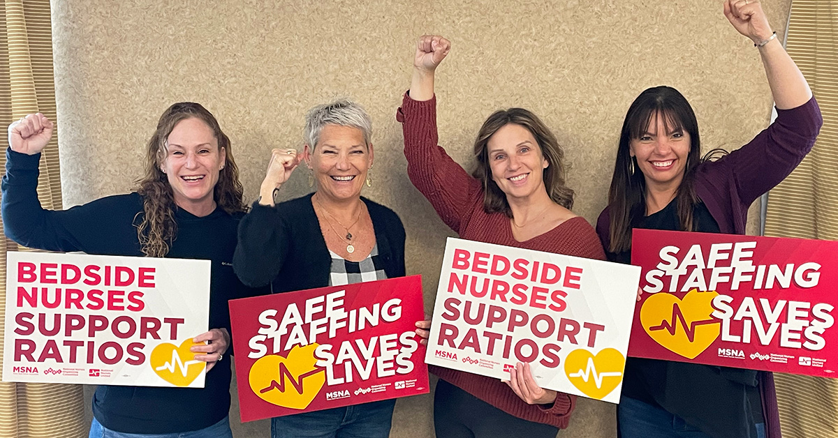 Four nurses with raisded fists hold signs in support of safe staffing ratios
