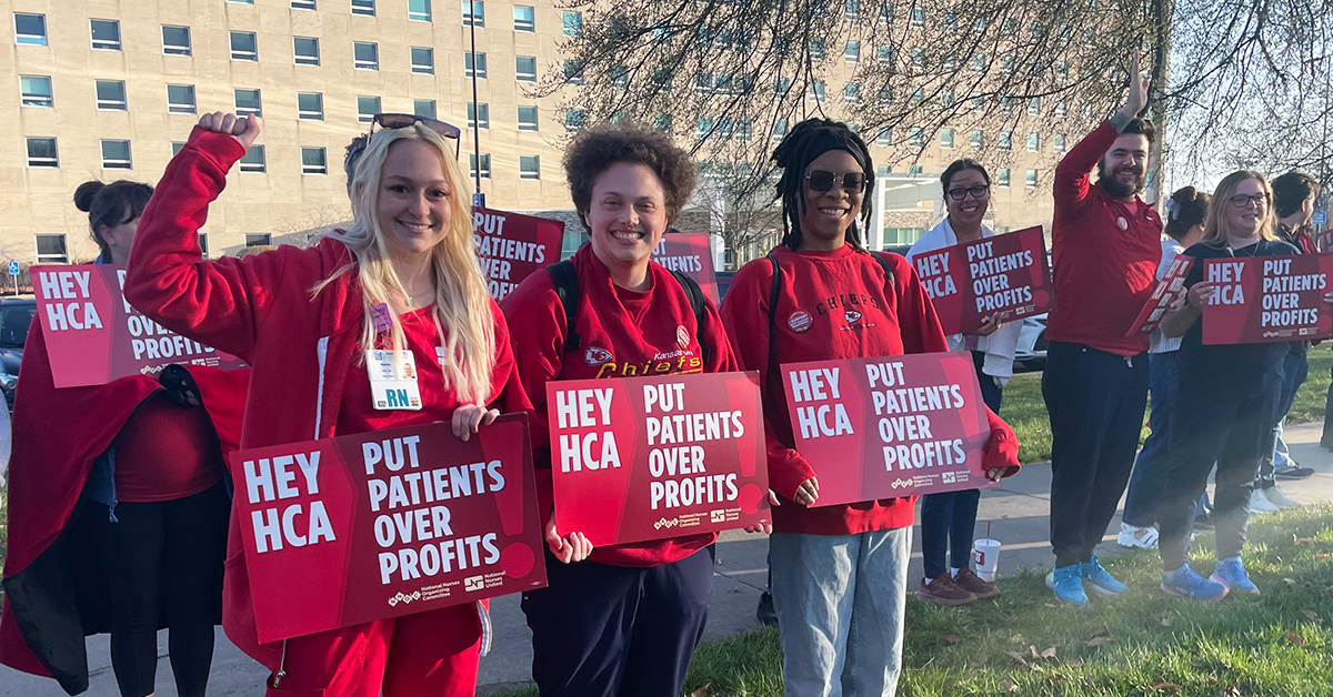Large group of nurses in blue scrubs outside hospital with raised fists holding signs "HCA Put Patients Over Profits"