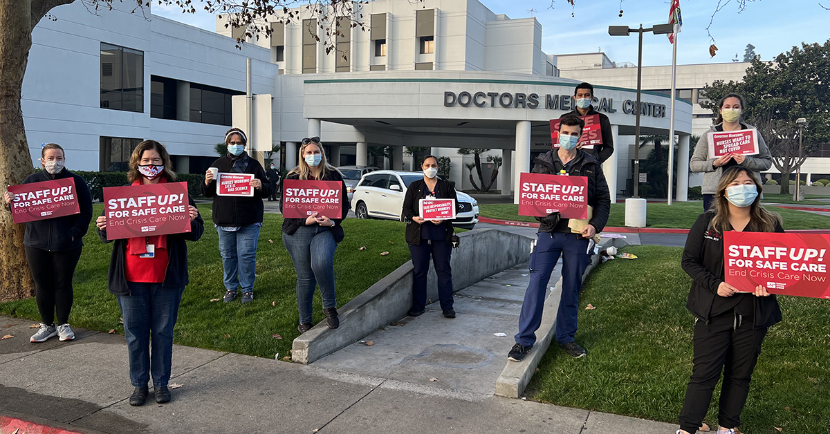 Nurses outside hospital with signs: "Staff up for safe care, end crisis care now"