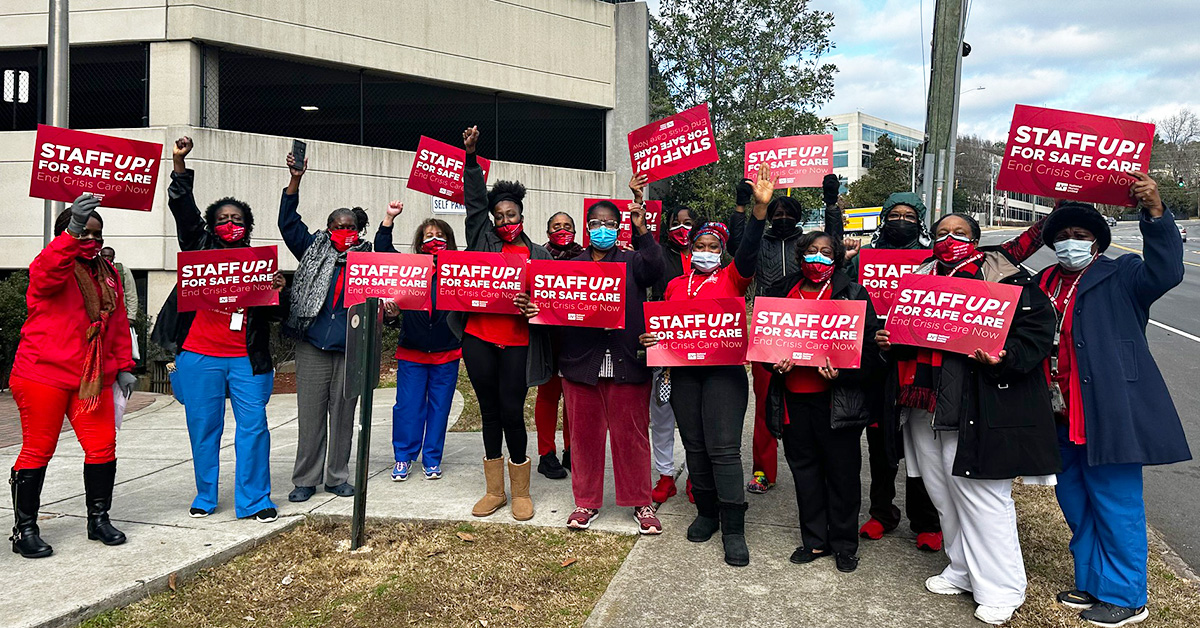 Large group of nurses outside hospital hold signs "Staff Up for Safe Patient Care"