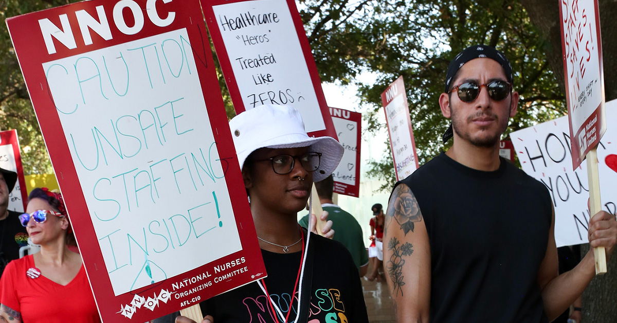 Nurses on picket line, one holds sign "Caution, Unsafe Staffing Inside"